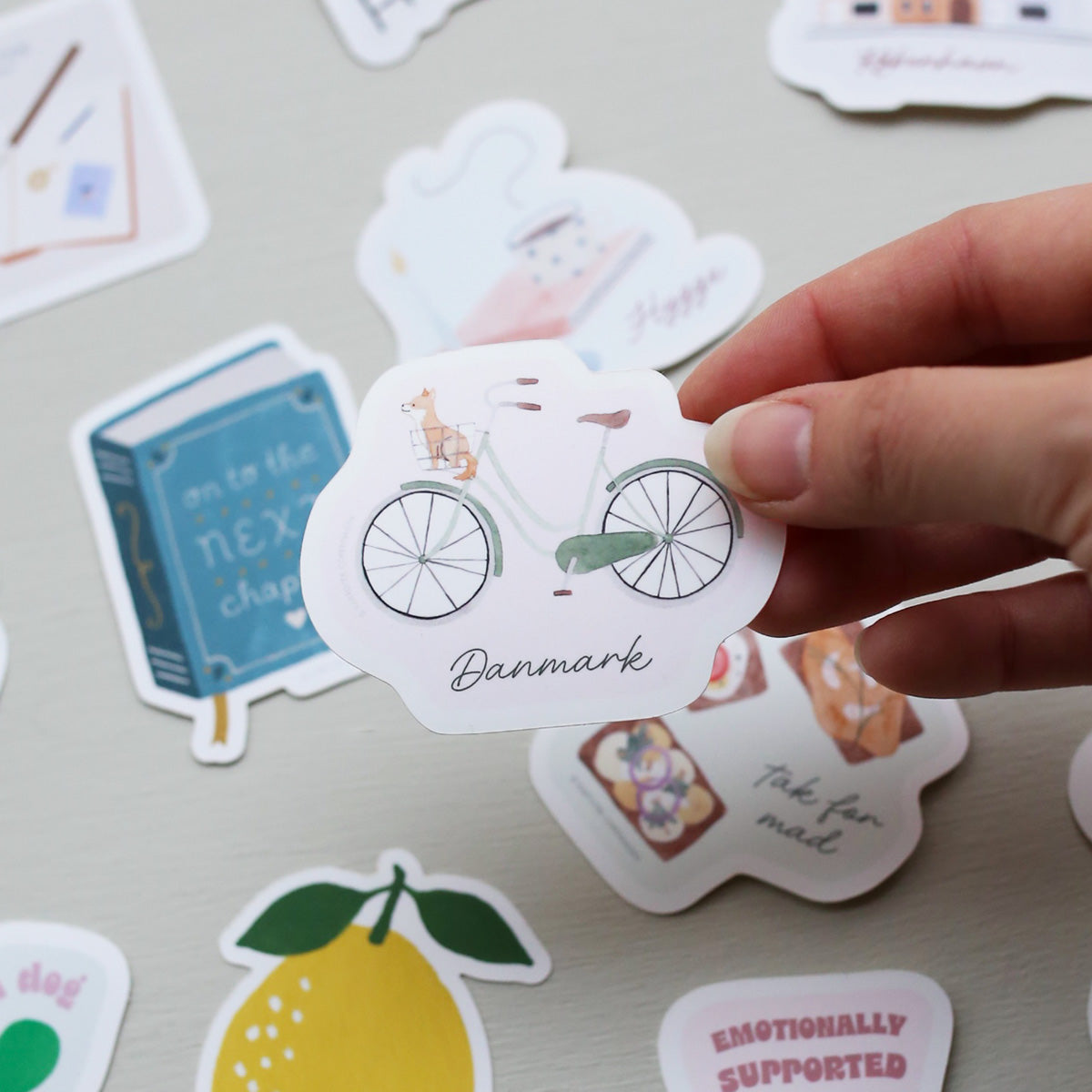 Hand holding a sticker with a bicycle design and 'Danmark' text on a surface with other stickers.