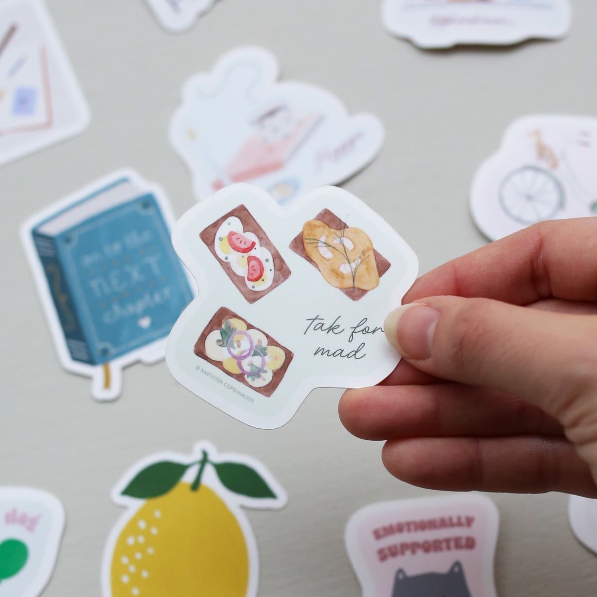 Hand holding a vinyl sticker with text and three Danish open-faced sandwiches called 'smørrebrød' on a light gray background with other stickers.