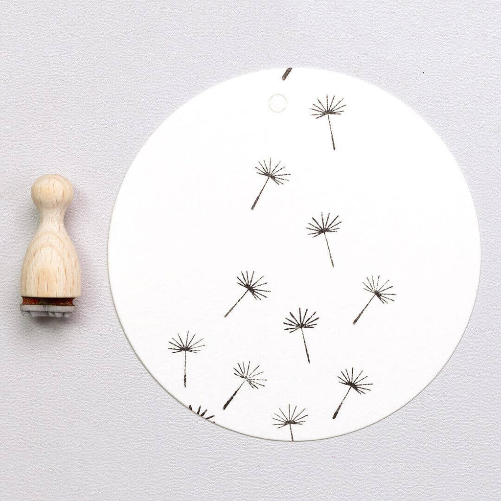 Round white board with black dandelion stamps and a wooden stamp on a light gray background