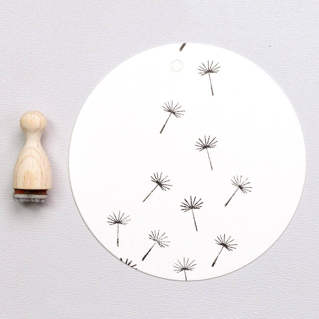 Round white board with black dandelion stamps and a wooden stamp on a light gray background