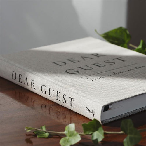 A hardcover book titled 'DEAR GUEST' with a textured light beige linen cover, resting on a dark wooden table with soft shadows and green ivy leaves.