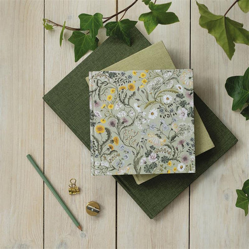 A floral-patterned fabric-covered notebook or journal with a gold-toned metal ring binder, styled on top of two dark green books with ivy leaves and a pencil on a light wood background.
