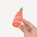 Hand holding a small bottle-shaped vinyl sticker with 'Certified Hot Mess' text on a light gray background