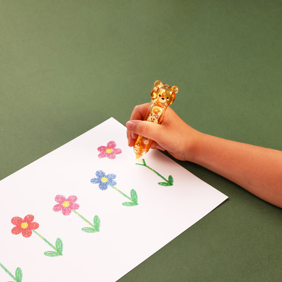 Hand holding a bear-shaped correction and glue tape container over a piece of paper with flower drawings on a green background