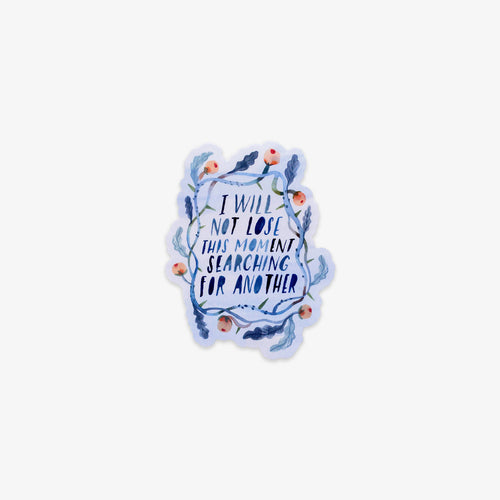 A die-cut sticker featuring the hand-lettered quote 'I WILL NOT LOSE THIS MOMENT SEARCHING FOR ANOTHER' in dark blue, surrounded by a watercolor-style floral wreath in shades of blue, purple, and peach, on a light gray background.