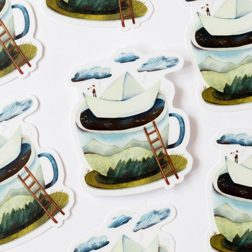 A close-up of repeated die-cut stickers showing a whimsical illustrated scene: a white paper boat floating on dark water inside a large blue-handled mug, with mountains, clouds, and tiny figures around it. The background is white.